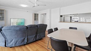 A modern living room with a gray sectional sofa, a white dining table with black chairs, a ceiling fan, and a kitchen area with appliances. - Front Row to the Tide at Imperial Surf (Gold Coast)
