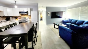 A modern kitchen with white cabinets, a dining table, and chairs. There is a blue sofa and a TV in the living area. - Blue Door House ! (Lakeland)