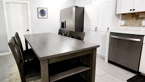 A modern kitchen with white cabinets, a stainless steel refrigerator, a dishwasher, and a dining table with chairs. - Blue Door House ! (Lakeland)