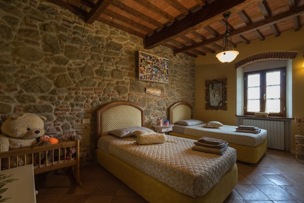 A room with two beds, stone walls, wooden beams, a large teddy bear, and a window with a view.