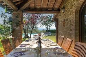 Un área de comedor al aire libre con una mesa dispuesta para seis personas, rodeada de muros de piedra y vistas a una piscina y árboles.