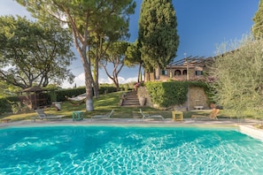 A swimming pool with lounge chairs, a stone building with a garden, and a tall tree.
