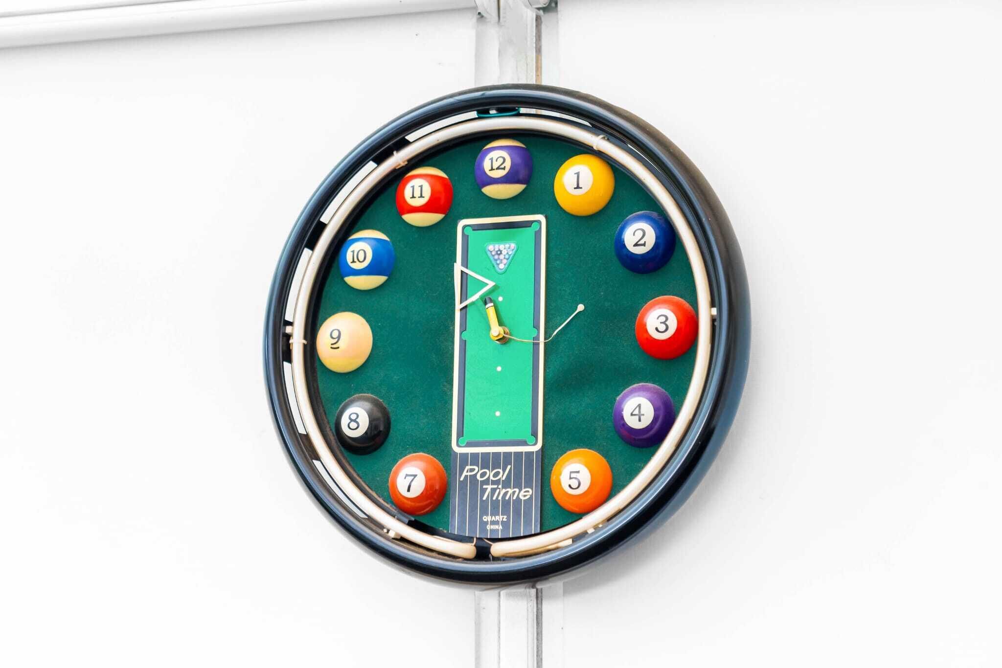 A wall clock with a pool table design, featuring numbered balls and a cue stick.