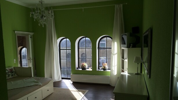A room with green walls, a white sofa, a small table with a vase, and three arched windows with curtains. - Vacation apartment in the tower  (Lehmkuhlen)