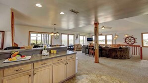 A spacious kitchen with a large island, wooden cabinets, and a fruit bowl. There is a dining area with a round table and chairs, and a cozy seating area with a large armchair and a wooden coffee table. - Oceanside, Ocean Views, Sound Views, Sundeck, Short Walk To The Beach! (Hatteras)