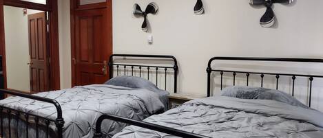 Une chambre avec deux lits, chacun doté d’un cadre en métal et d’une literie grise. On y trouve trois ventilateurs de plafond avec des pales noires et des détails argentés. Une porte en bois avec un panneau en verre est visible à l’arrière-plan.