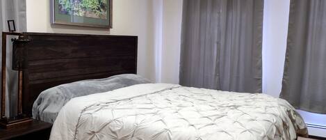 A bedroom with a wooden headboard, a bed with a white quilt, a painting of flowers on the wall, and grey curtains.
