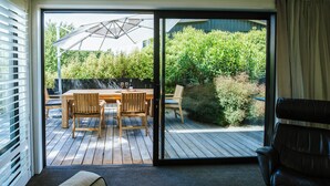 Quarto com porta de vidro deslizante que dá para um alpendre externo com mesa e cadeiras de madeira, guarda-sol e vista para um jardim.