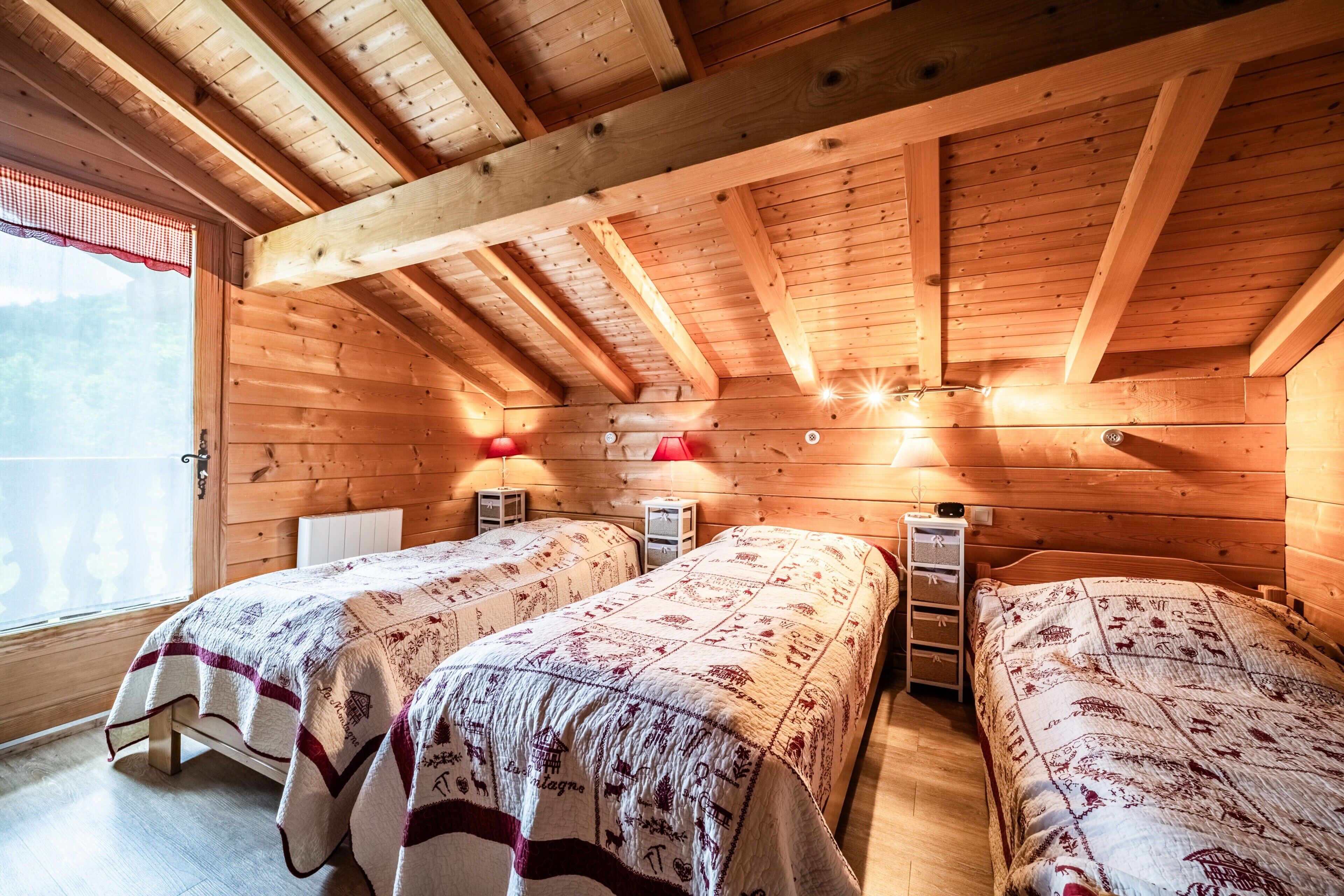 A wooden cabin with two beds, a window with a red curtain, and a wooden ceiling.