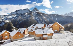 Un conjunto de cabañas de madera con tejados nevados, frente a un paisaje de montañas nevadas y un cielo despejado.