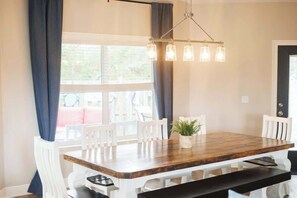A dining area with a wooden table, white chairs, a hanging light fixture, and a window with curtains.