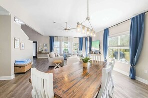 A spacious dining area with a wooden table and white chairs, a ceiling fan, and large windows with blue curtains.