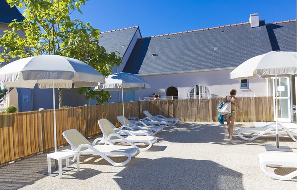 A poolside area with sun loungers and umbrellas, a wooden fence, and a building with a tiled roof in the background.