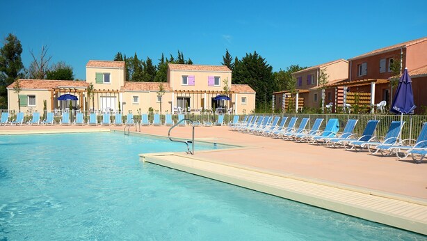 Una piscina con sillas de descanso y sombrillas, rodeada de edificios de estilo mediterráneo.