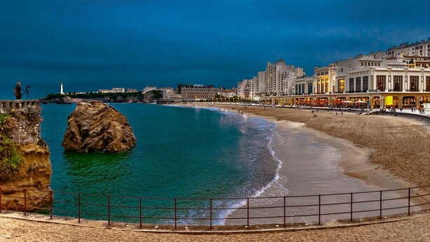 Escena costanera amb un promontori rocos, una platja sorrenca i un passeig marítim amb edificis.