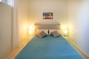 Ein Schlafzimmer mit einem Bett, zwei Nachttischlampen, einem Wandbild und einem Fenster mit Vorhängen.
