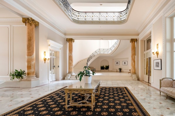 Eine beeindruckende Lobby mit Marmorboden, einem zentralen Tisch und einer Treppe zu den oberen Stockwerken.