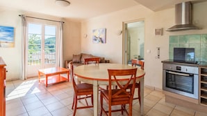 A dining area with a round table and chairs, a sofa, a kitchen with a stove, and a view through a glass door. - Charmant T4 climatisé | Accès aux piscines (Callian)