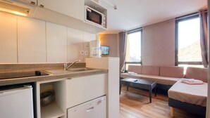 Une cuisine compacte équipée d’un four à micro-ondes, d’un évier et d’un petit réfrigérateur. Le coin salon comprend un canapé et une table basse, et la chambre est dotée d’un lit avec un oreiller.