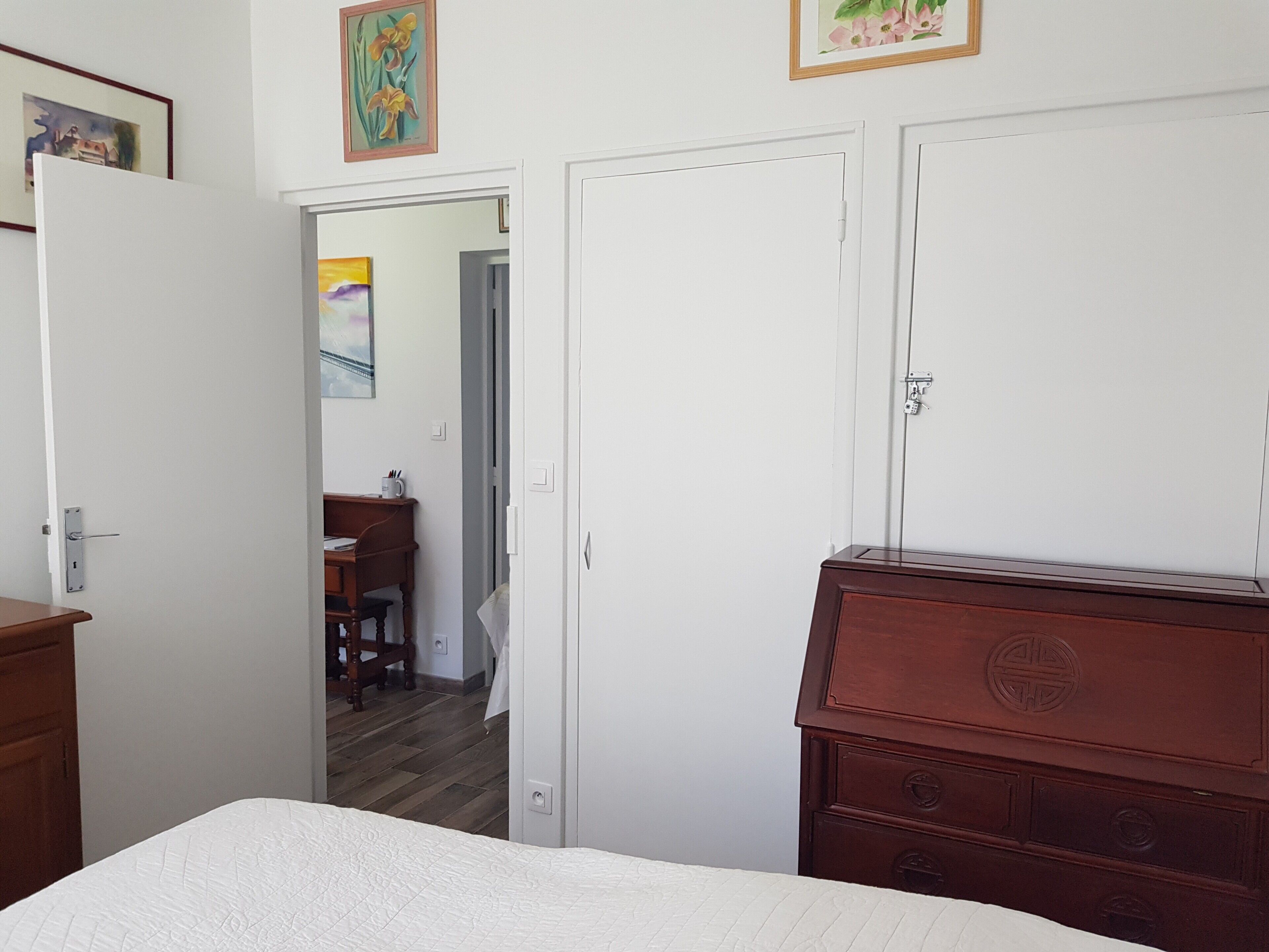 A bedroom with a bed, a wooden dresser, a desk with a chair, and a door leading to another room.