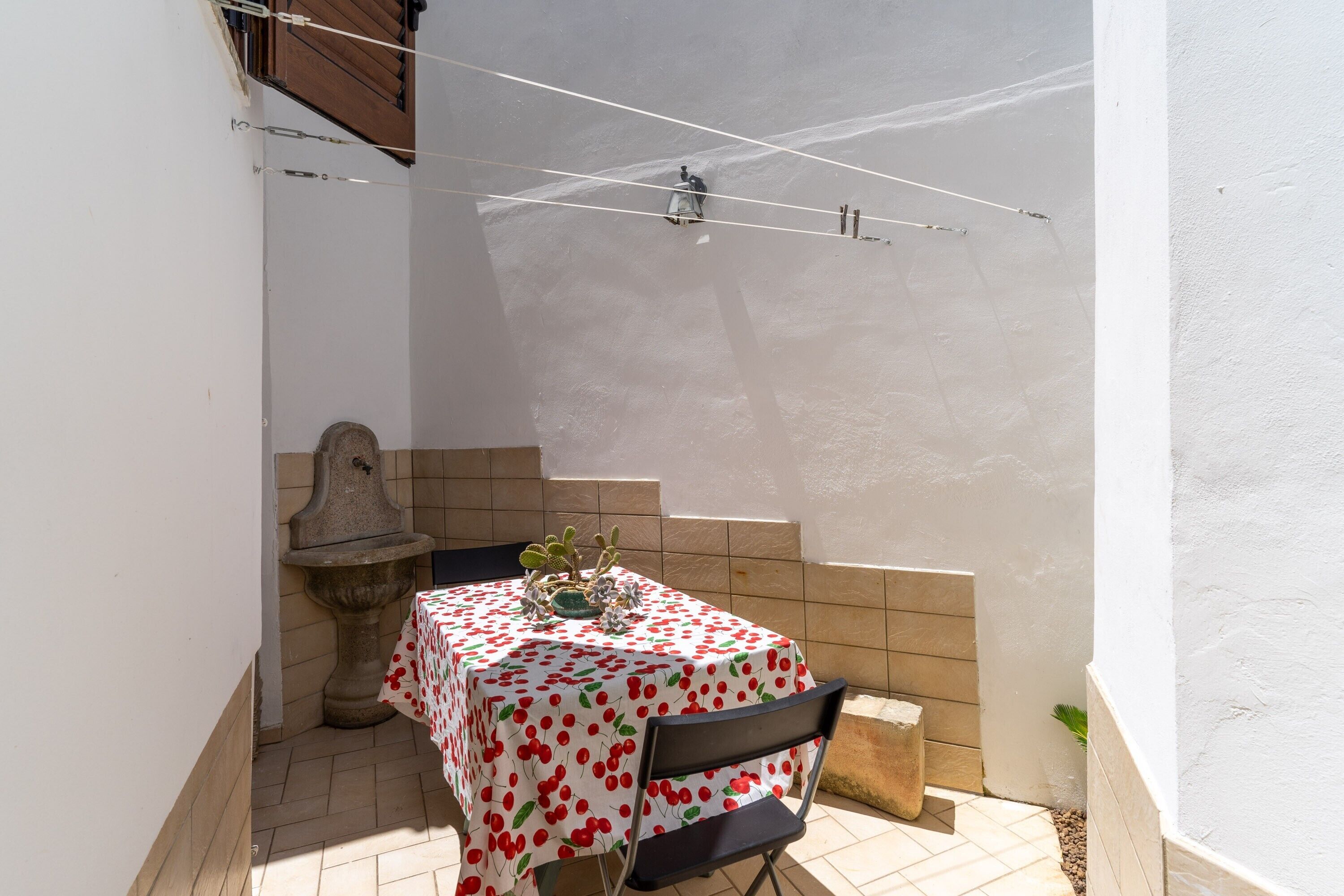 Un pequeño patio con una mesa cubierta con mantel estampado de cerezas, una silla, una maceta de piedra y un muro de azulejos.