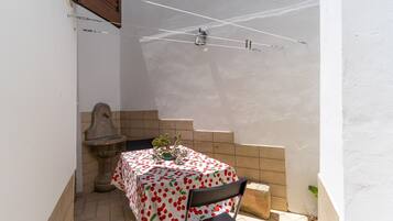 Un pequeño patio con una mesa cubierta con mantel estampado de cerezas, una silla, una maceta de piedra y un muro de azulejos.