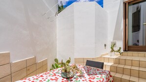 Een terras met een tafel, gedekt met een tafelkleed met kersenprint, een cactus in een schaal en een plant in een pot.