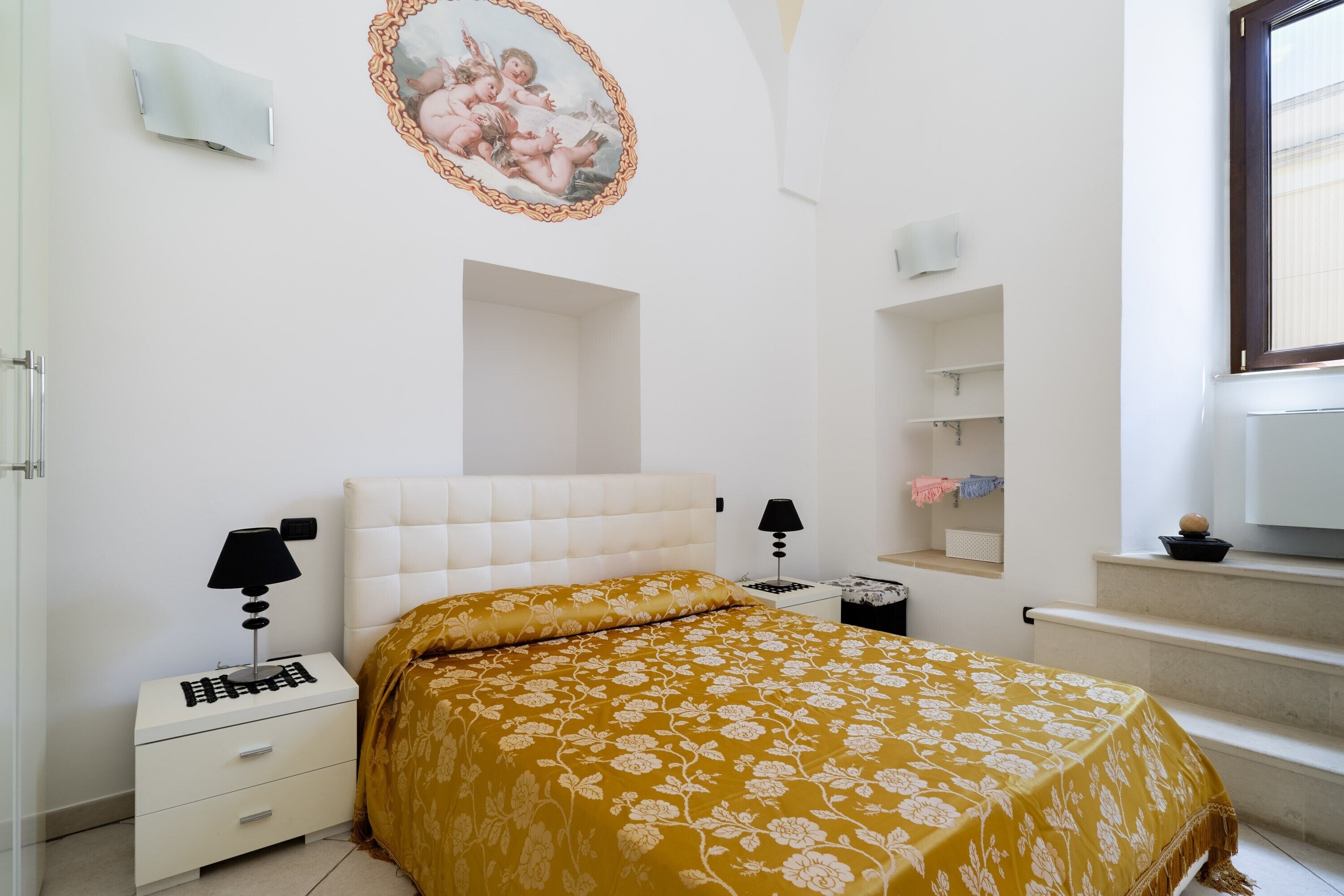 A bedroom with a bed, nightstands, a lamp, a wall-mounted shelf, and a window.