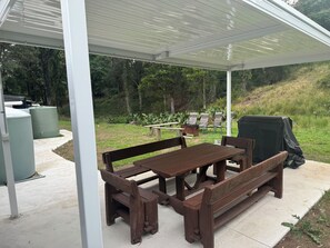 Un espace extérieur abrité, doté d’une table de pique-nique et de bancs, entouré de verdure et d’arbres.