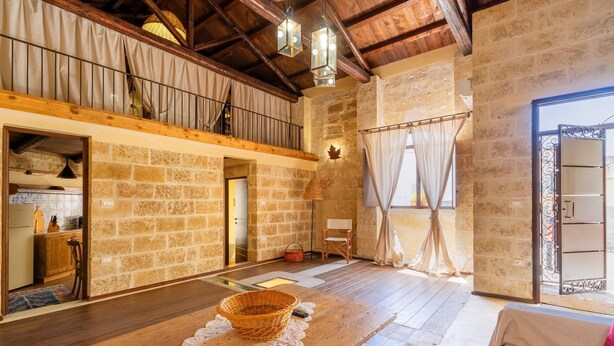 Amplia habitación tipo loft con vigas de madera a la vista, una pared de piedra, una mesa de madera con una cesta, un sillón acogedor y vistas a través de un ventanal.