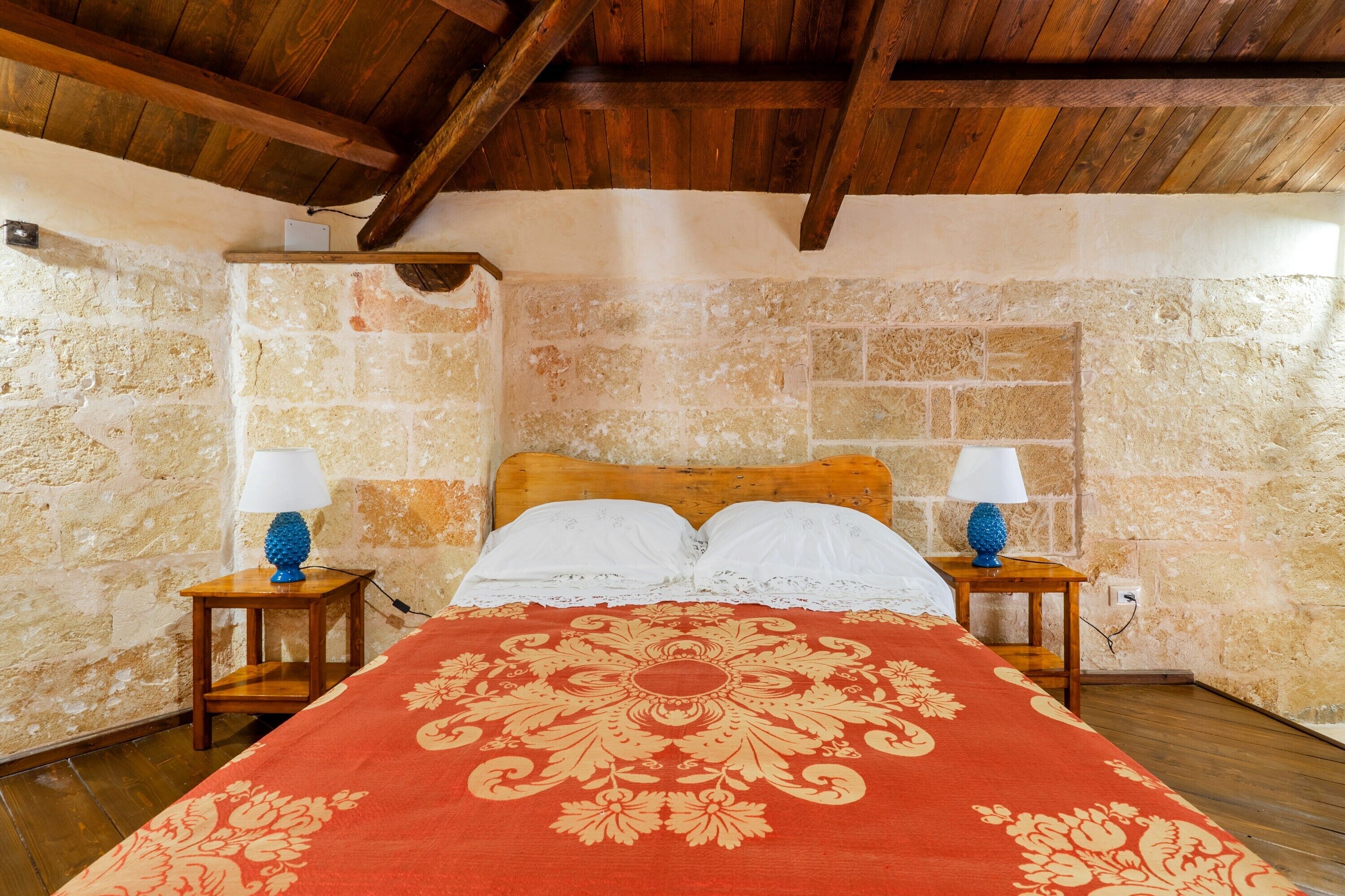 Une chambre avec un mur en pierre, des poutres apparentes en bois au plafond, un lit recouvert d’une couvre-lit rouge et or, deux tables de chevet avec des lampes et une tête de lit en bois.