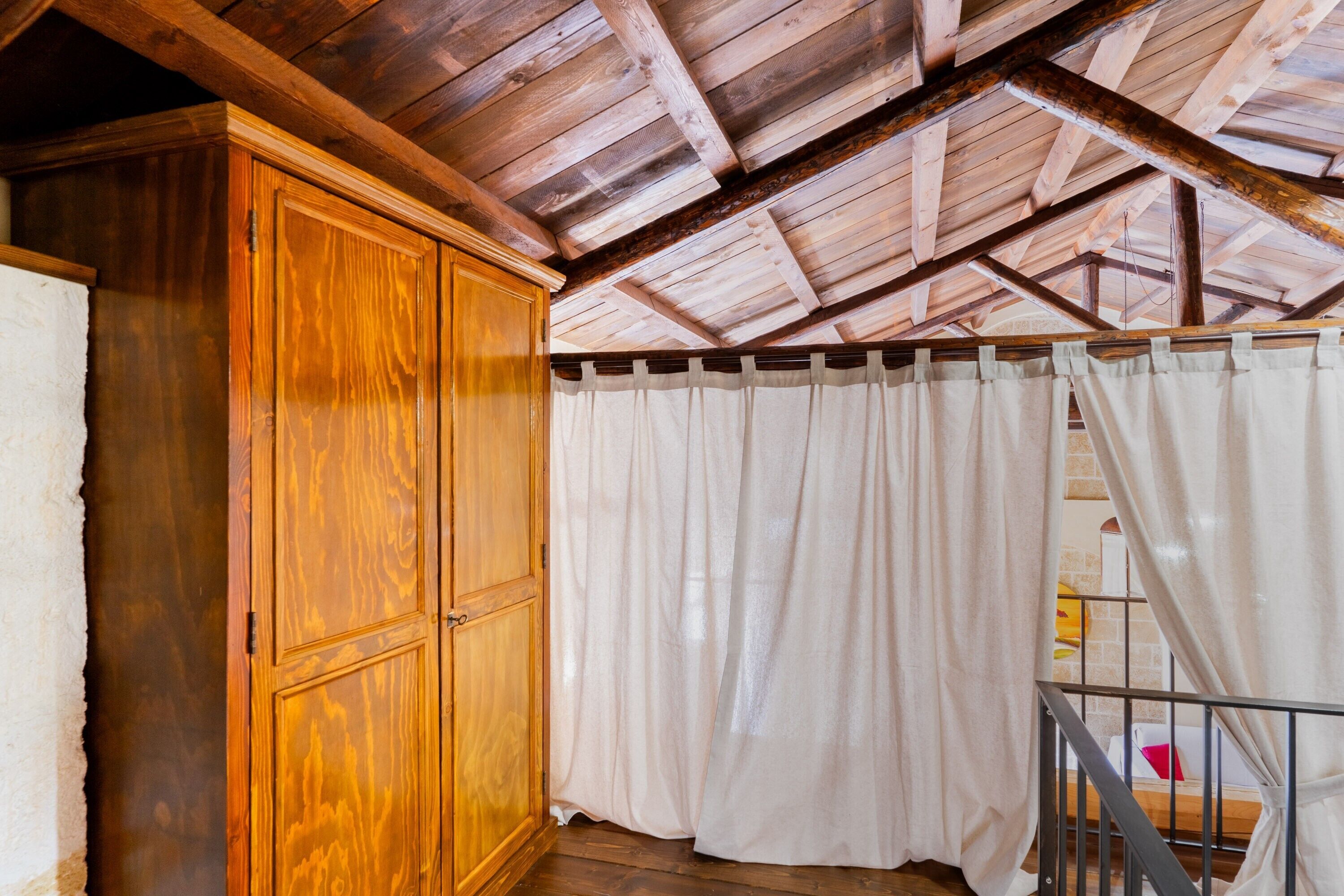 A room with wooden beams on the ceiling, a wooden wardrobe, and white curtains.