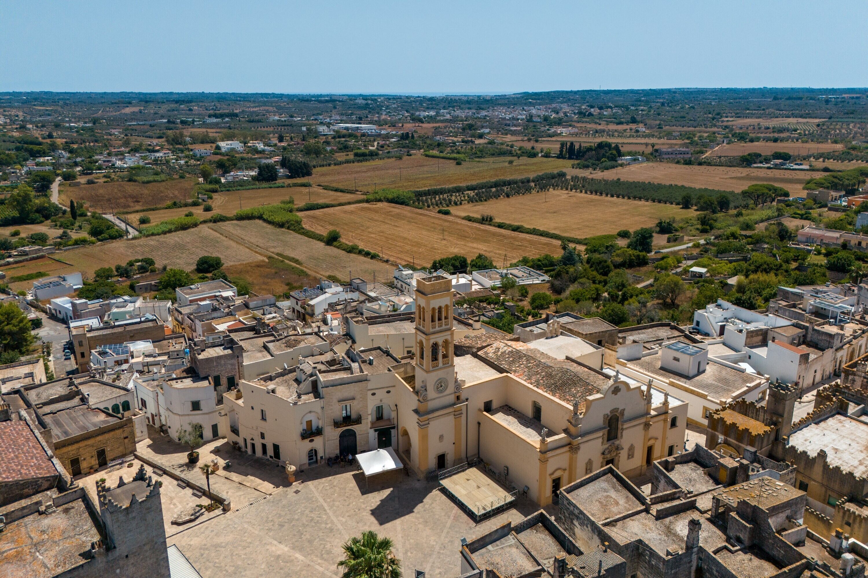 An aerial view of a town with a prominent church featuring a bell tower, surrounded by residential buildings and agricultural fields.