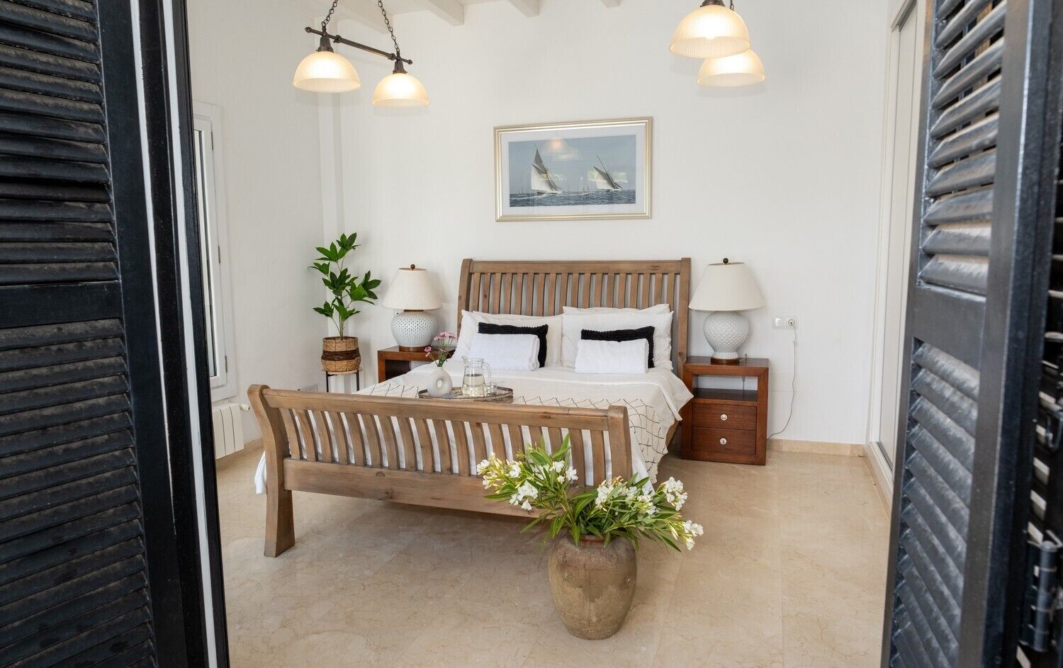 A bedroom with a wooden bed, bedside tables with lamps, a potted plant, and a framed picture on the wall.