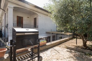 Un barbecue installé dans un espace extérieur, avec un bâtiment blanc, un arbre et un jardin clôturé.