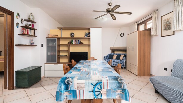 Een eethoek met een tafel, bedekt met een tafelkleed met een nautisch thema, een plafondventilator en een bankstel op de achtergrond.