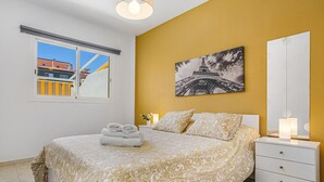 A bedroom with a bed, a nightstand, a wall-mounted lamp, a painting of the Eiffel Tower, and a window with a view of buildings.