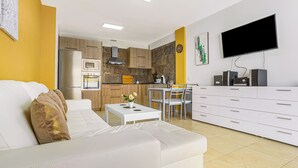 A modern living room with a white sofa, a dining area with a table and chairs, a kitchen with wooden cabinets and a built-in oven, and a mounted TV.