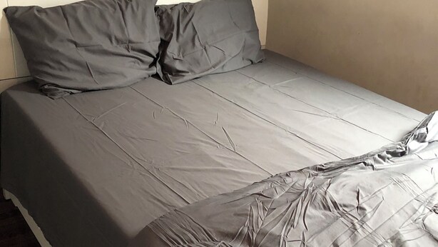 Una cama con ropa de cama gris, un cabecero blanco y una mesita de noche con una lámpara.