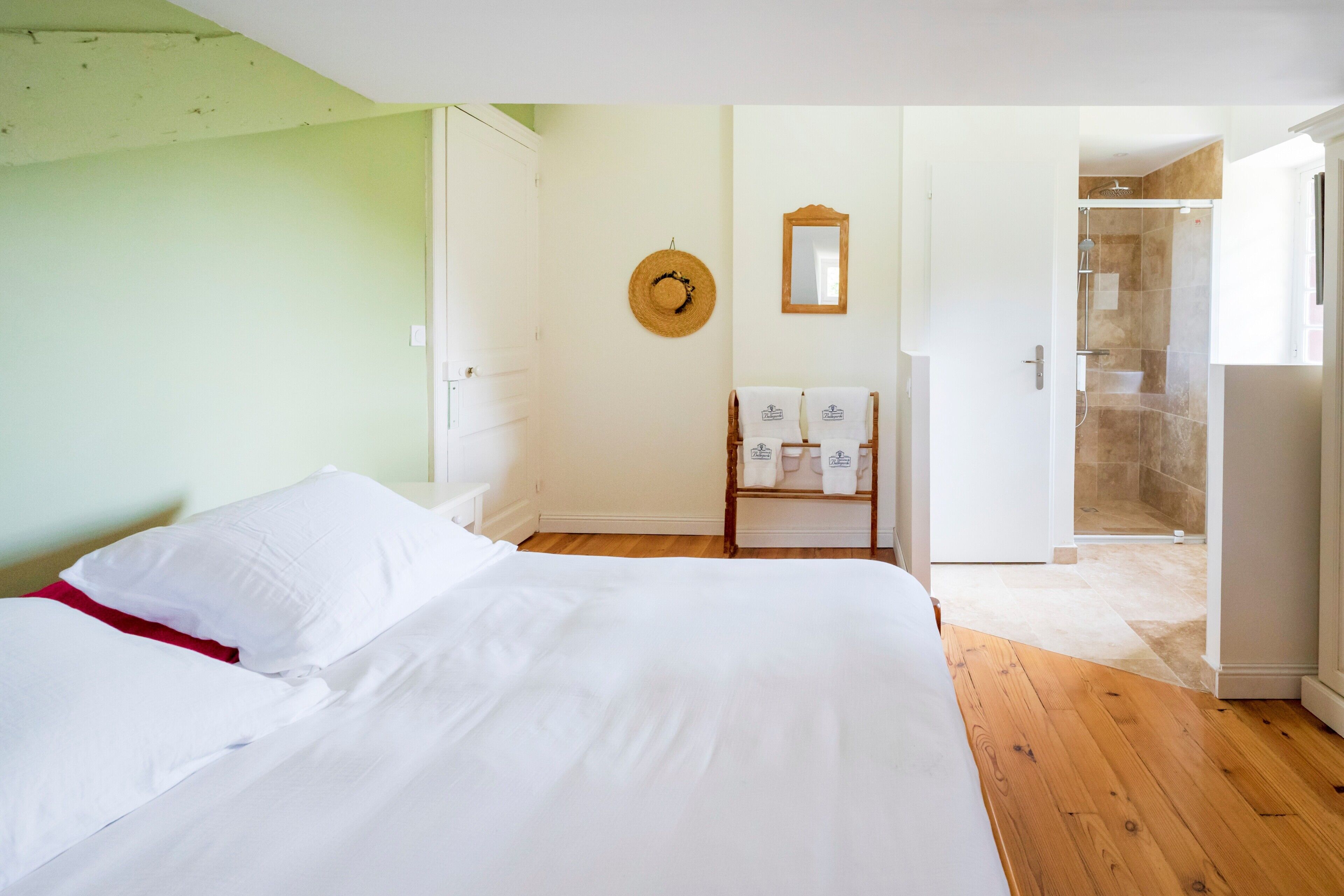 A hotel room with a bed, a wooden floor, a shower area, and a wall-mounted hat.