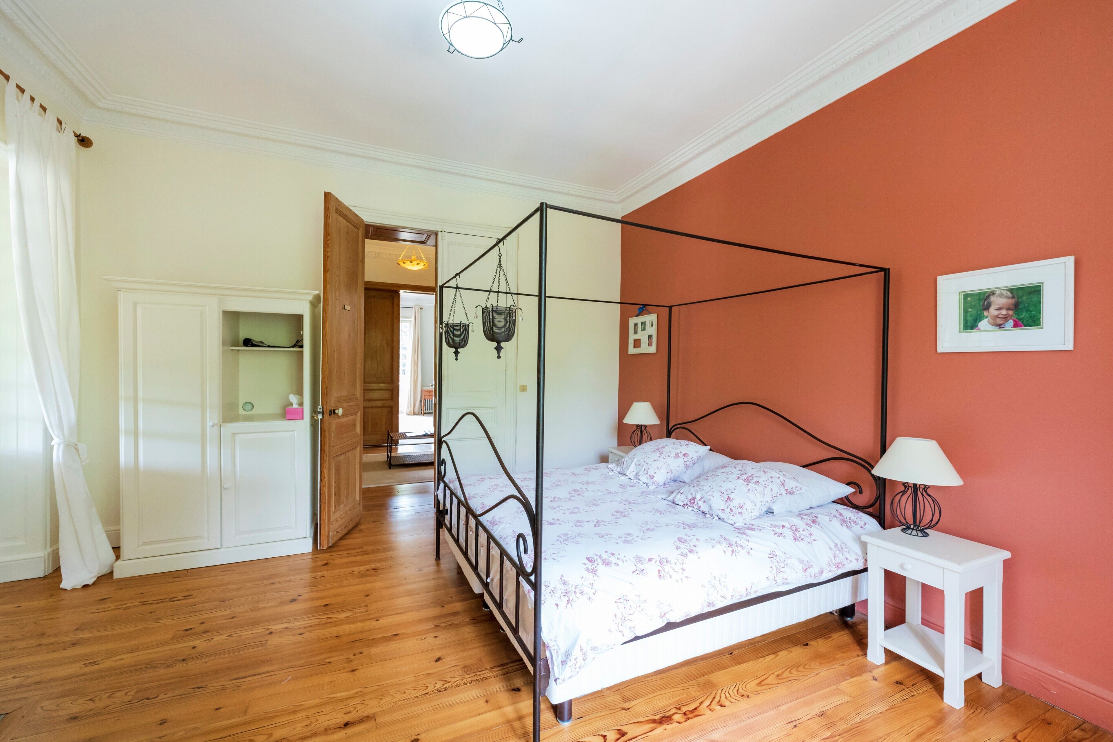 Un dormitorio con cama con dosel, una mesita de noche con lámpara, un armario y una puerta que da a otra habitación.
