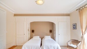 Una habitación con dos camas, un sillón de mimbre y una puerta blanca.