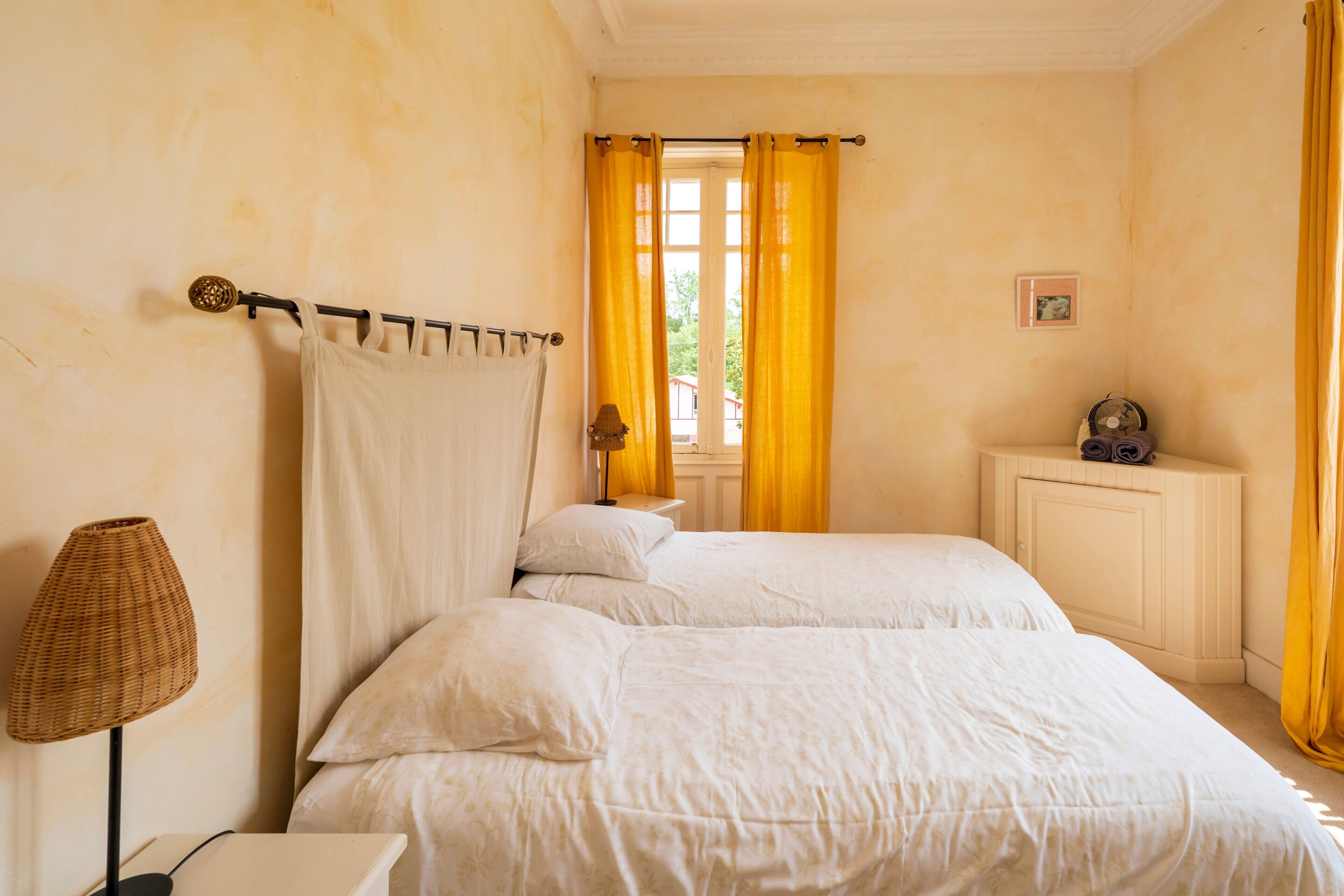 A room with two beds, a bedside table with a lamp, a small cabinet, and a window with yellow curtains.