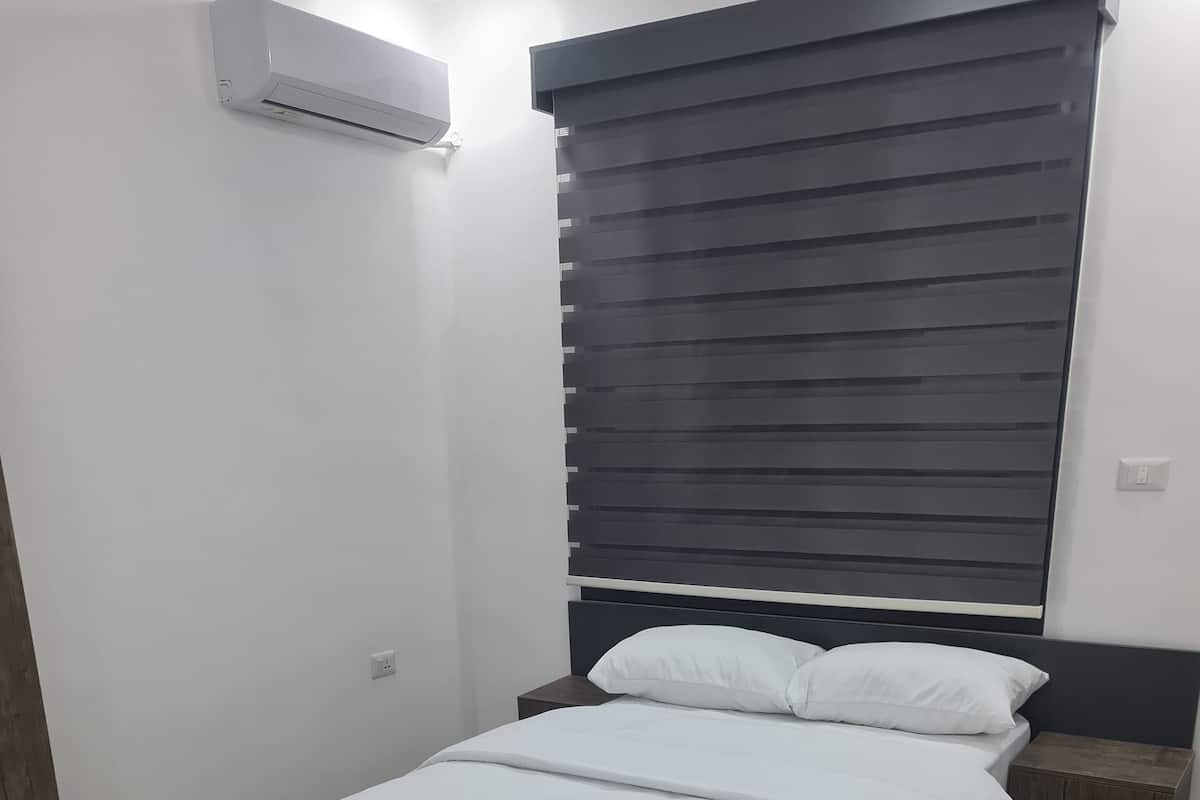 Une chambre à coucher comprenant un lit, une table de chevet, un climatiseur fixé au mur et une fenêtre avec des stores.