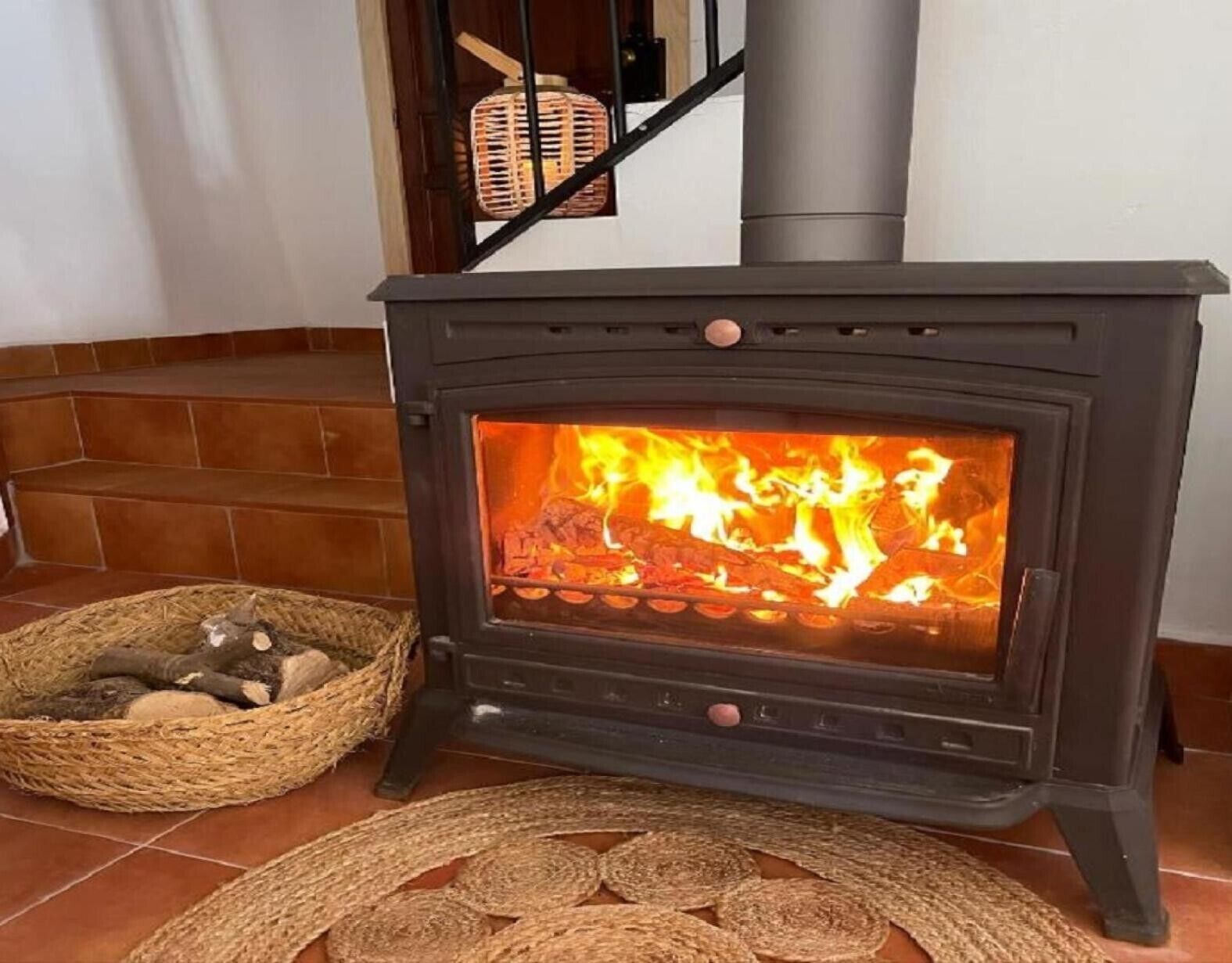 Ein Kaminofen mit Feuer, ein geflochtener Korb voller Holzscheite und eine gewebte Matte auf dem Boden.