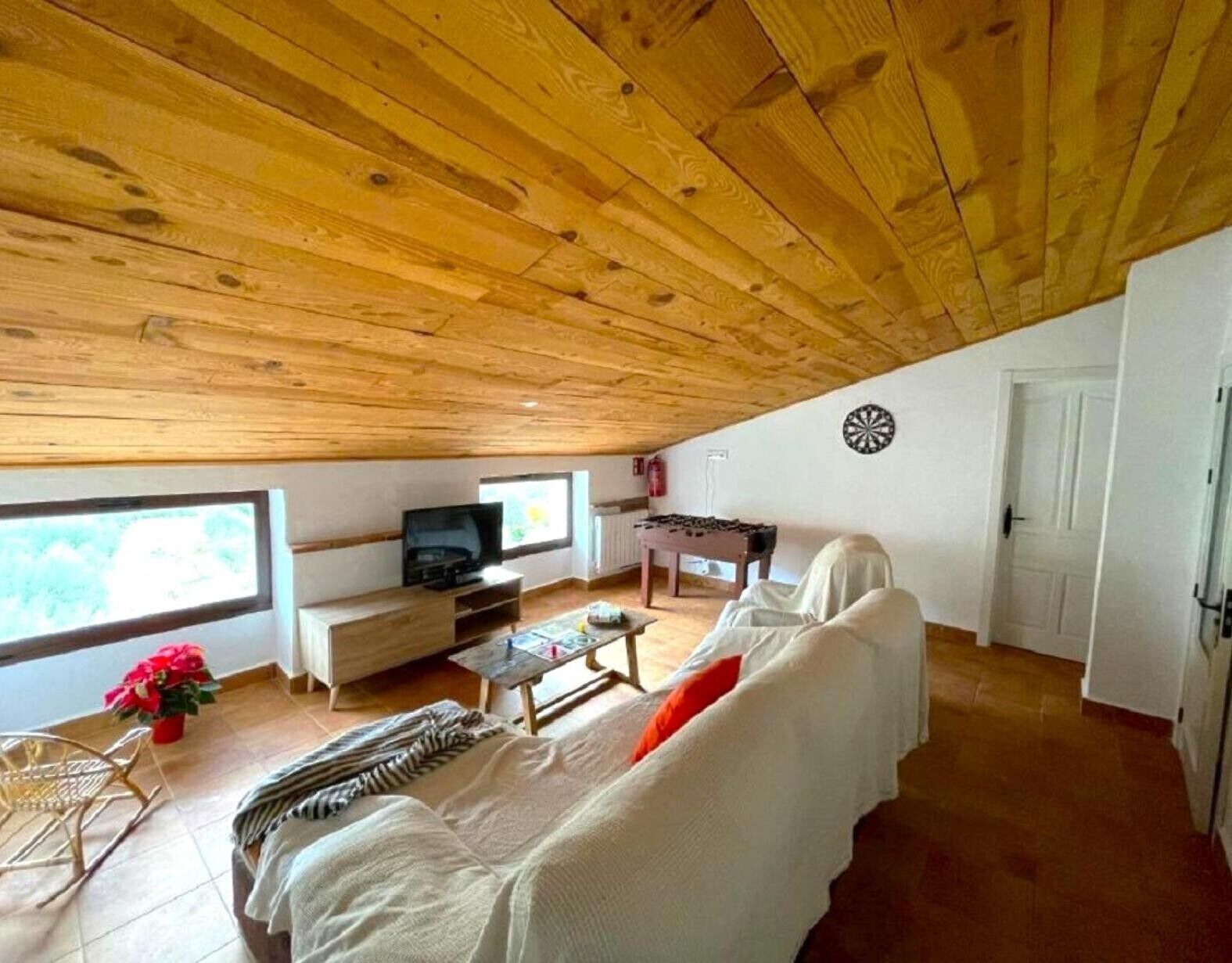 A cozy, wooden-paneled room with a sofa, a television, a dartboard, and a window.