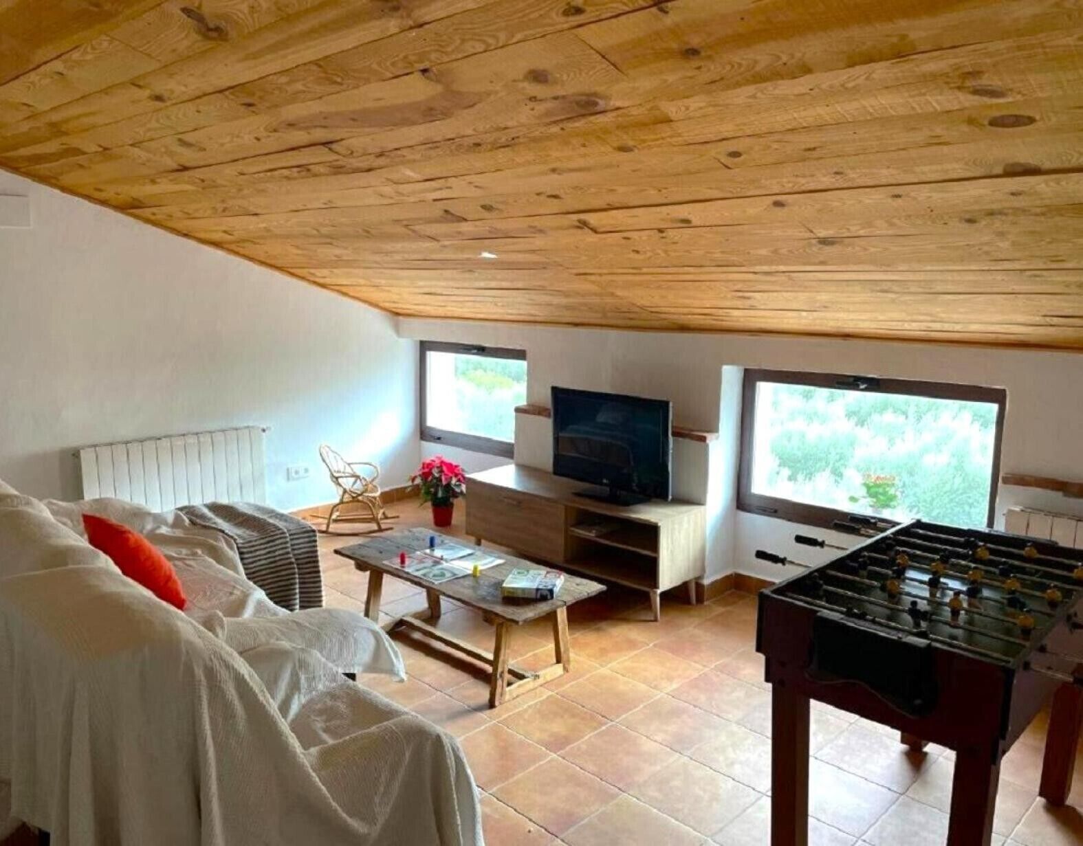 Un salon chaleureux avec un plafond en bois, un canapé, un téléviseur, une table de baby-foot et une fenêtre donnant sur un beau paysage.