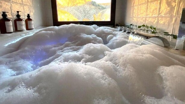 A large, clear bathtub filled with bubbles, a wooden ceiling, and a window with a view of a landscape.