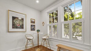 A room with white walls, a wooden floor, two bar stools, a round table, a bench, and large windows with shutters.