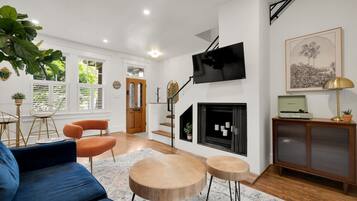 A modern living room with a blue sofa, wooden coffee table, orange chairs, a TV, a fireplace, and a large plant.